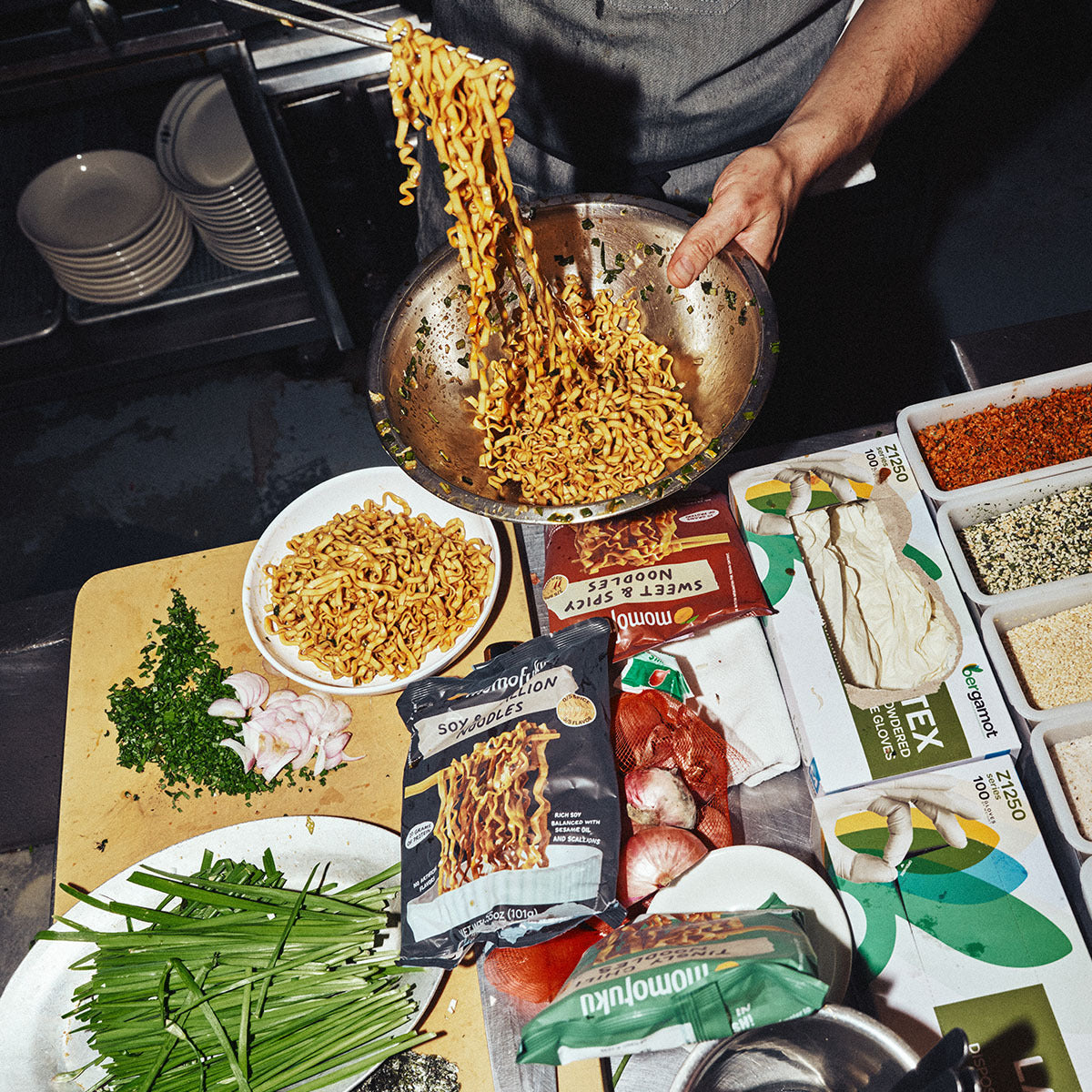 A person holds a large metal bowl filled with stir-fried noodles above a spread of ingredients and packaged food on a dark kitchen counter. Visible items include a package labeled 'Soyumaki', a bowl of chopped green onions, parsley, garlic, and various seasoning containers.