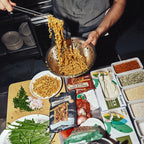 Person cooking noodles in a pan with various ingredients and packages on a table.