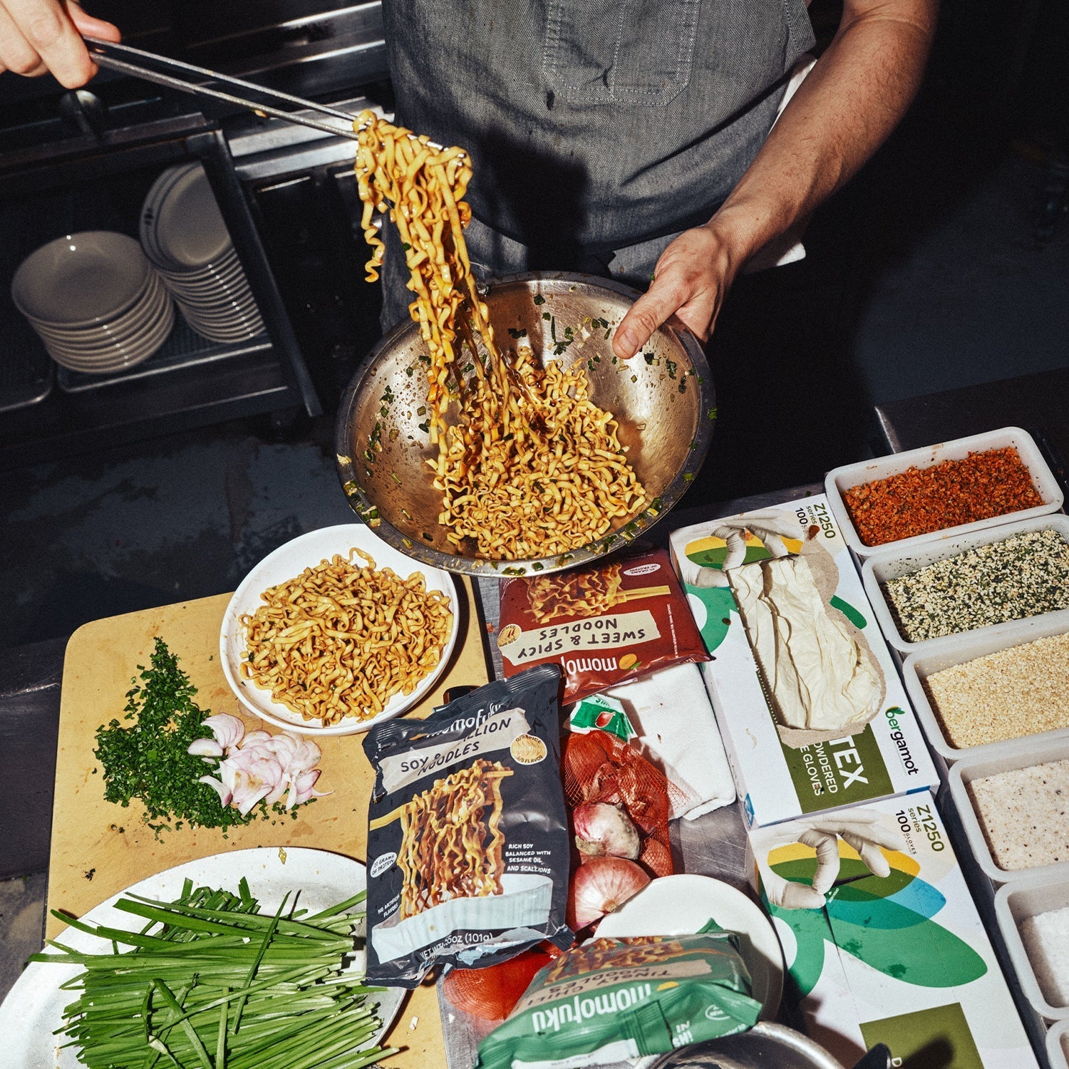 Person cooking noodles in a pan with various ingredients and packages on a table.